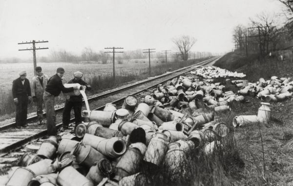 A group of four men standing on a railroad tracks, dump milk onto the ground from a milk can. Around them are hundreds of empty milk cans strewn about the ground.