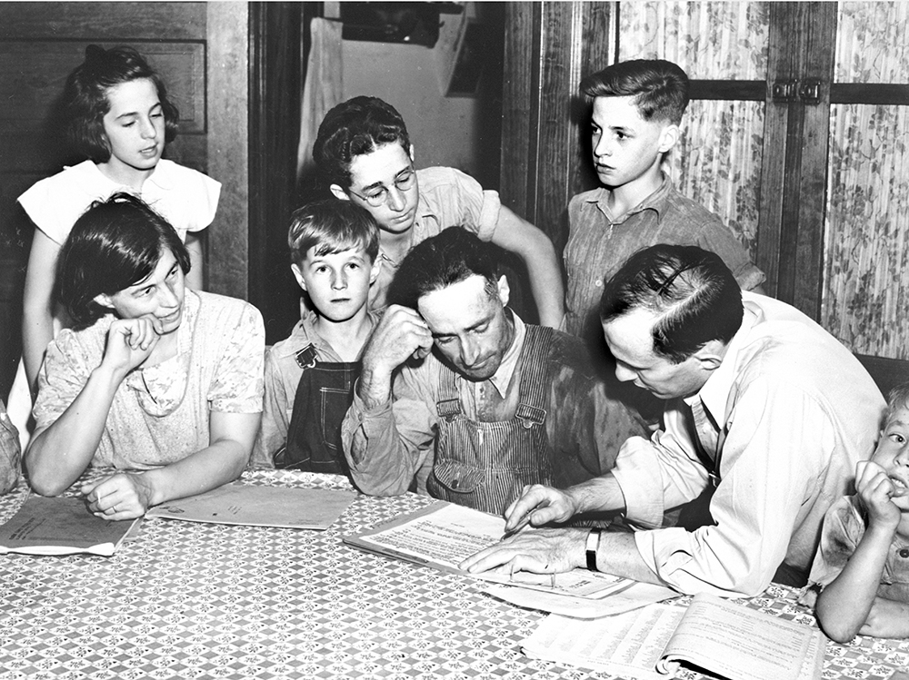 Two men and a woman sit at a table pouring over a document. Children crowd around.