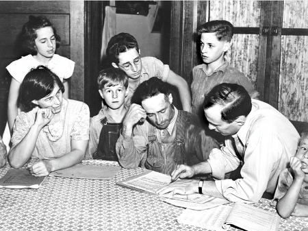 Two men and a woman sit at a table pouring over a document. Children crowd around.
