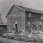 Black and white photograph of potters standing in front of the Pauline Pottery complex. There are two buildings: the larger workshop is a two-story building, and behind it is a one-story kiln house with a bottle kiln emerging from the roof.