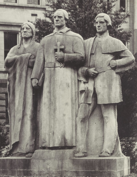 Statue of three figures. At the center is a man in religious vestments, to his left is a man in European clothing, and to his right is a Native American figure.