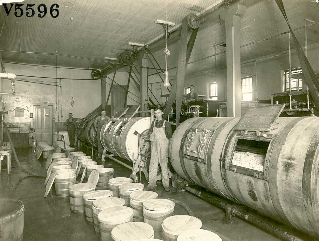 Two men stand next to industrial butter churns at a creamery.