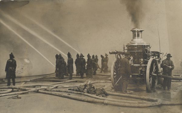 A small group of fire fighters fight an unseen fire with three fire hoses powered by a steam-powered pump. Smoke obscures the fire, and on the right, a fire engine has smoke billowing from its steam engine.