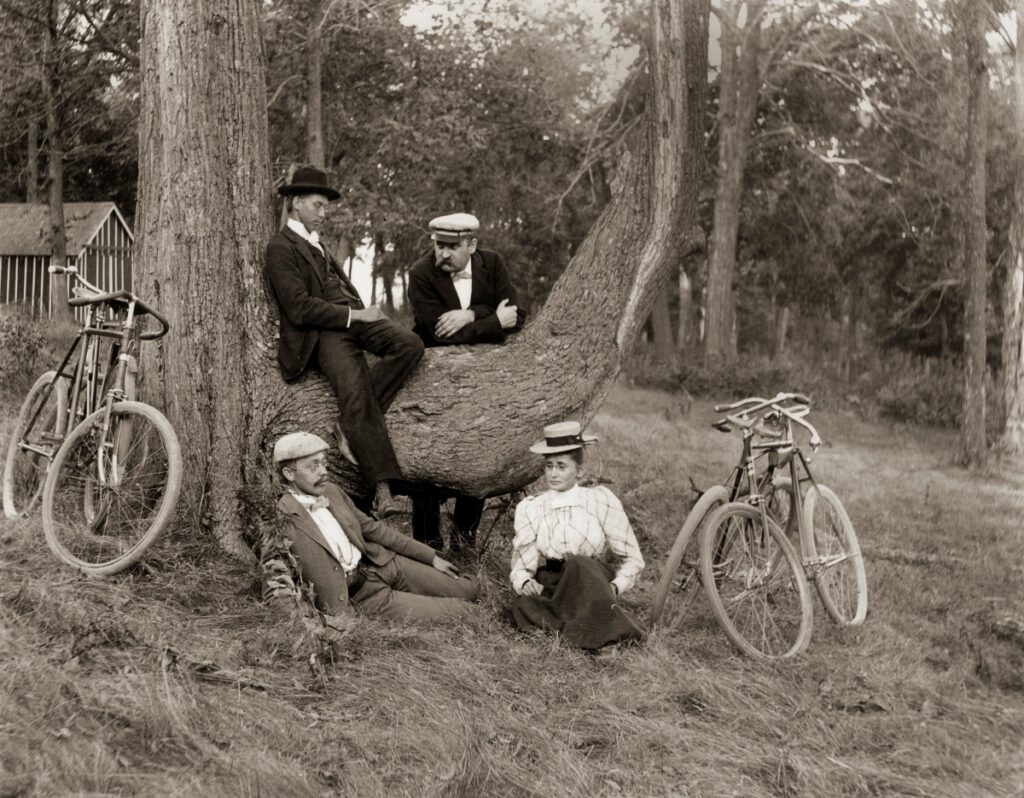 A group of three men and one woman pose in rest against a tree. Their bicycles nearby.