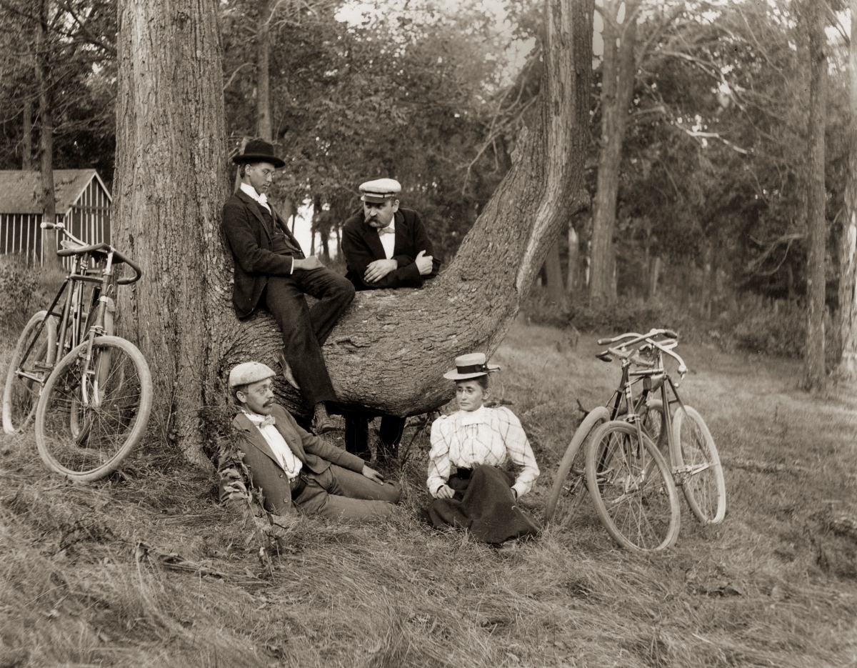 A group of three men and one woman pose in rest against a tree. Their bicycles nearby.