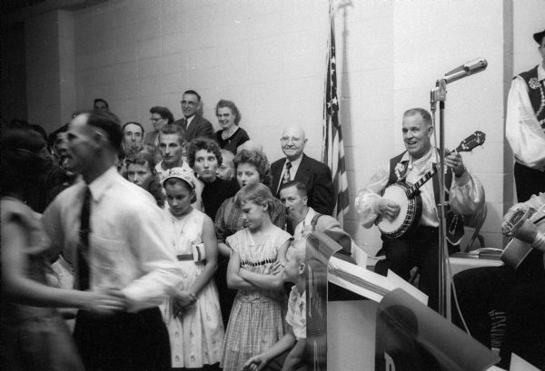 A crowd gathers to watch men and women dance the polka at the Verona Legion Hall. A man plays a banjo in the background.