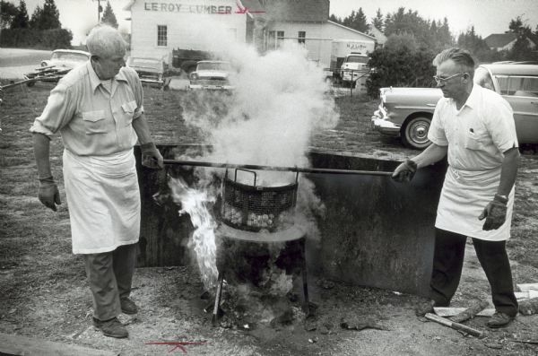 Two men use a iron bar to lift a basket from a boil pot. Both men wear protective gloves and aprons.