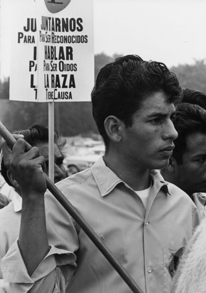 An Obreros Unidos (United Workers) member holding a sign, possibly like those in the background, that says "Juntarnos Para Ser Reconocidos/Hablar Para Ser Oidos/La Raza Tiene Causa"