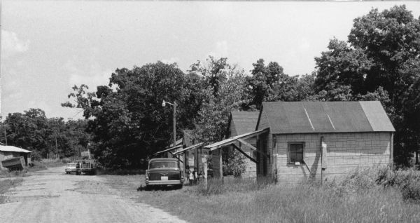 A row of small wooden framed buildings with front porches on posts. The buildings are faced with asphalt siding. At the center, three children are standing in front of a building beside a black car looking into the camera. Farther down the road on the left is a shack and a worker standing by a car across from it.