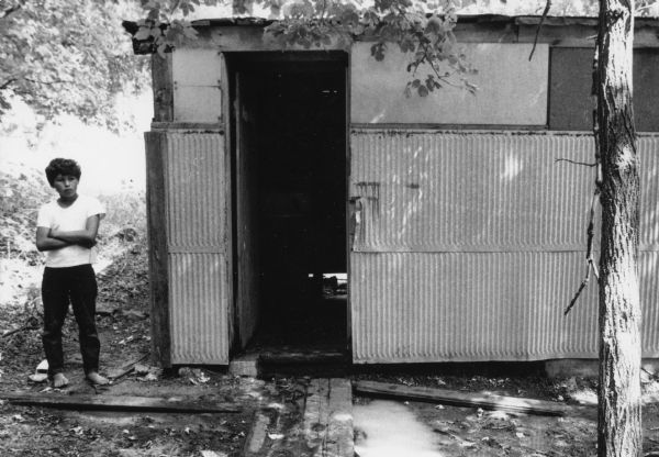 A boy wearing a white short-sleeved t-shirt and dark pants is barefoot and standing to the left of a communal shower hut for migrant farm workers in a Wautoma labor camp. The communal hut is floorless and has a tin exterior.