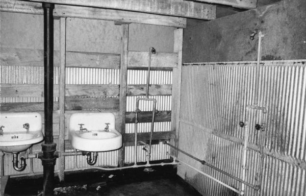 The interior of a floorless tin communal shower hut with shower heads and sinks for migrant farm workers in a labor camp.