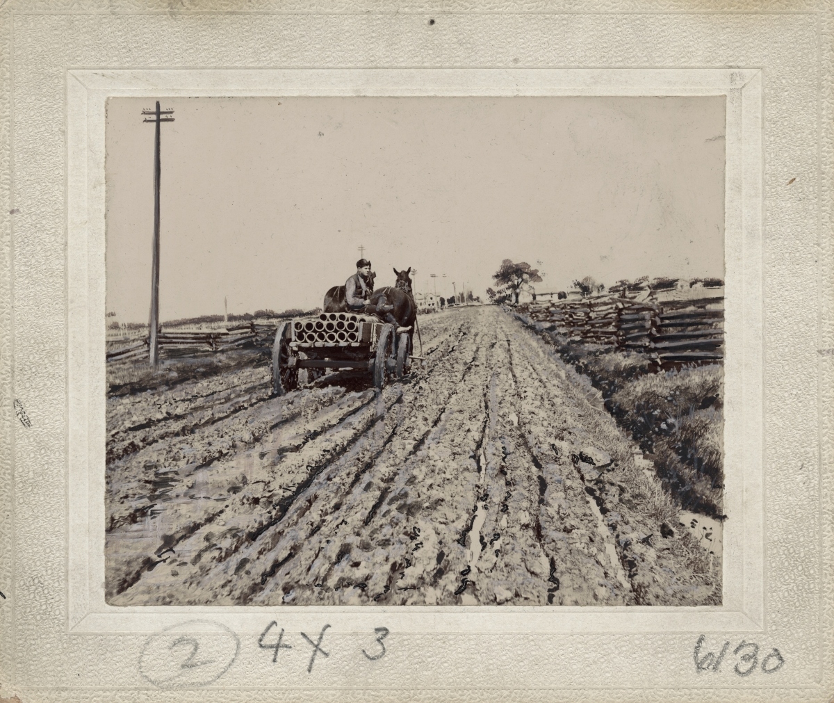 Rear view of a man driving a horse-drawn wagon filled with pipe on a muddy, rutted dirt road. A split-rail fence can be seen on both sides of the road and a telephone pole appears on the left. In the far distance are a farm house, buildings and trees.
