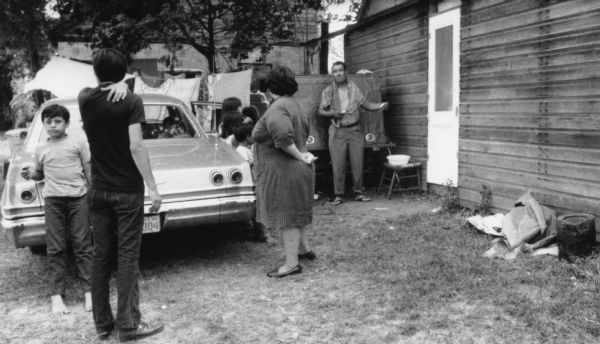 A migrant worker family stands in front of a parked vehicle and a cabin that serves as temporary housing in a Wautoma labor camp. Some of the children are not wearing shoes. The shirtless man with a towel around his neck is washing or shaving himself with water in a small basin beside him.