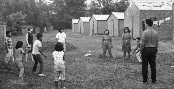 A group of children stand in a circle playing a ball game in a labor camp. Behind them is a line of identical small buildings.