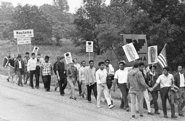 A group of about 30 men march alongside a highway past the Wautoma City Limits sign. They carry signs some with the image of the Lady of Guadelupe, others with the National Farm Workers Association banner with the Aztec eagle symbol.