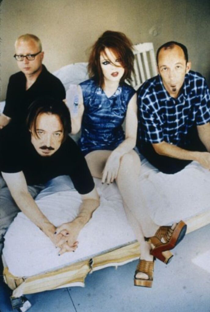Color publicity photograph of the band members from Garbage, a Madison, Wisconsin rock band. They are sitting or leaning on a bed with white linens.