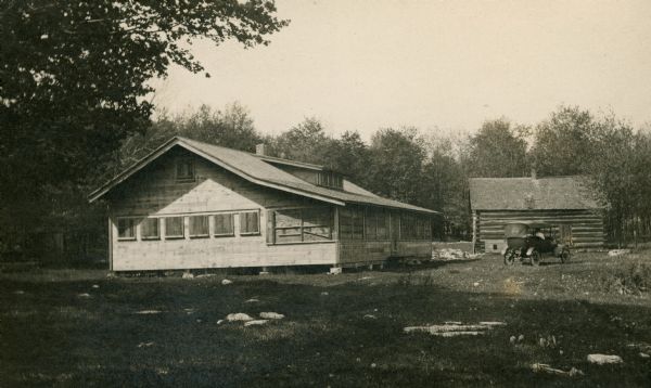 A black-and-white photograph of two buildings at a cherry camp. One building is the camp house with a full-span porch, shuttered, and the other is a log cabin.
