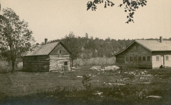 A black-and-white photograph of two buildings at a cherry camp, orchard in bloom visible in the background. One building is a log cabin and the other is a wood frame camp house with all of its windows boarded up.