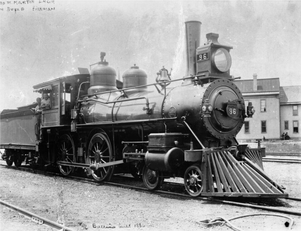Black and white photograph of a train locomotive, the engineer poses out the side of the cab window.