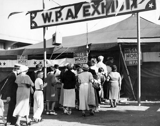 A photograph of a line of people waiting to enter a large tent. A sign above them reads W.P.A Exhibit.