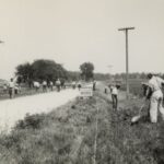 A black and white photo of men digging out a ditch on either side of the road, at the center of the image is a sign that reads "USA WORK PROGRESS WPA"