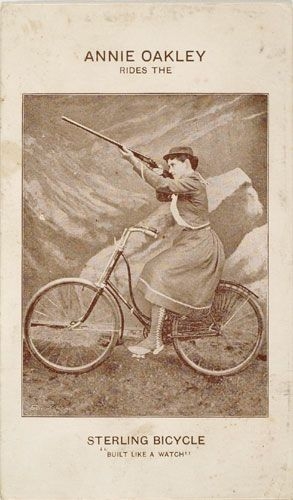 A photo advertisement of Annie Oakley, in a dress astride a Sterling bicycle against a painted backdrop. She aims a rifle into the air. Text copy reads, “Annie Oakley rides the Sterlin Bicycle. Built like a watch.”