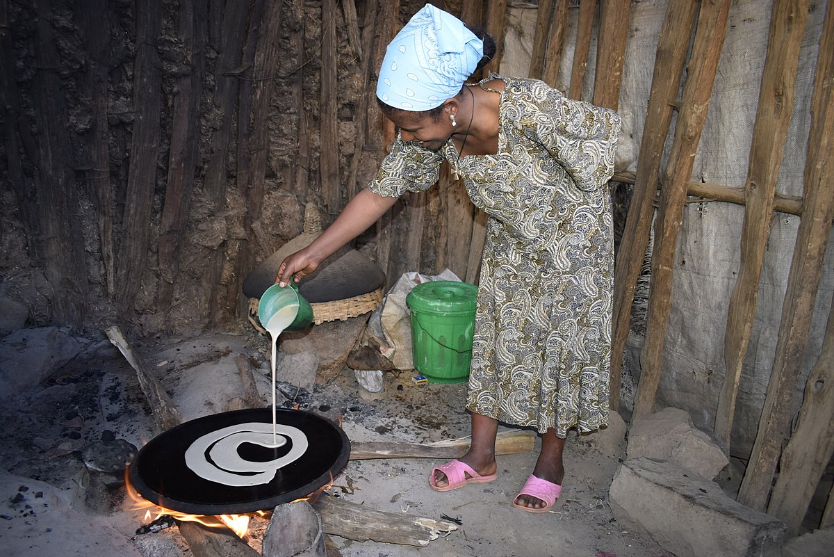 A photograph of a woman pouring batter on a griddle over an open fire, making Injera in Ethopia.