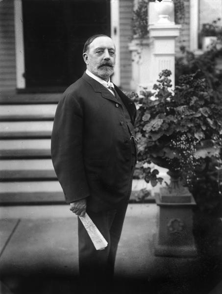 Photo portrait of William Horlick in front of his house holding a rolled newspaper.