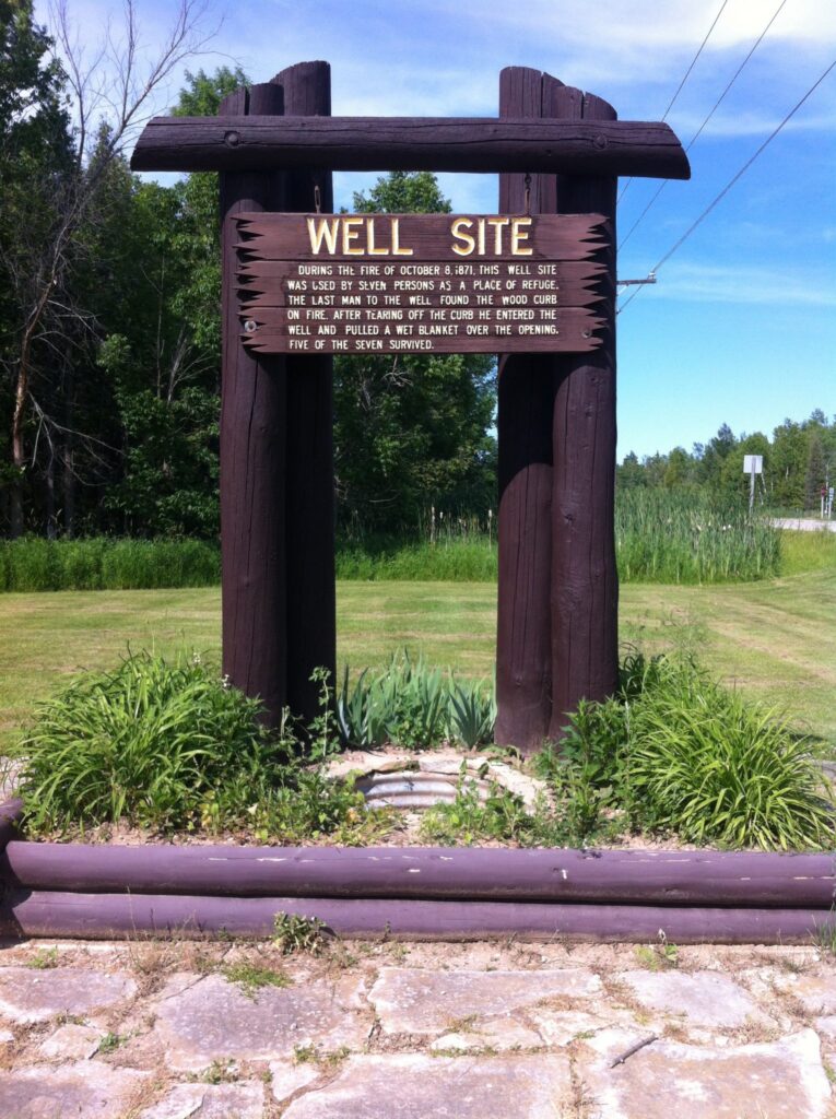 Photograph of a historical marker over a well that reads "Well Site. During the fire of October 8, 1871, this well site was used by seven persons as a place of refuge. The last man to the well found the wood curb on fire. After tearing off the curb he entered the well and pulled a wet blanket over the opening. Five of the seven survived.