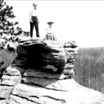 A black-and-white photograph of a man and a woman posed atop a rock formation.