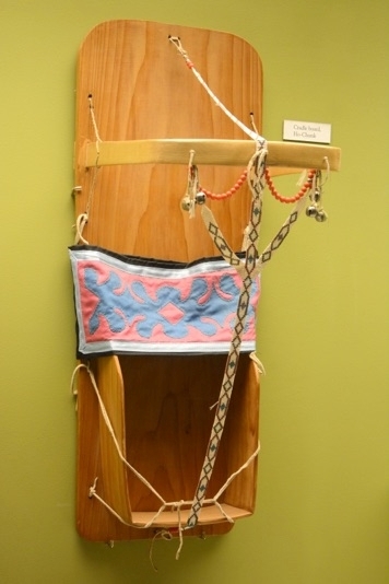 A cradleboard: a wooden board that a infant would lay on, with sidewalls at the bottom where the child's feet would go. There is a textile band around the center and cords to contain the child, and a bentwood handle. beaded straps connect the handle to the bottom of the piece.