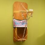 A cradleboard: a wooden board that a infant would lay on, with sidewalls at the bottom where the child's feet would go. There is a textile band around the center and cords to contain the child, and a bentwood handle. beaded straps connect the handle to the bottom of the piece.