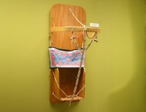 A cradleboard: a wooden board that a infant would lay on, with sidewalls at the bottom where the child's feet would go. There is a textile band around the center and cords to contain the child, and a bentwood handle. beaded straps connect the handle to the bottom of the piece.