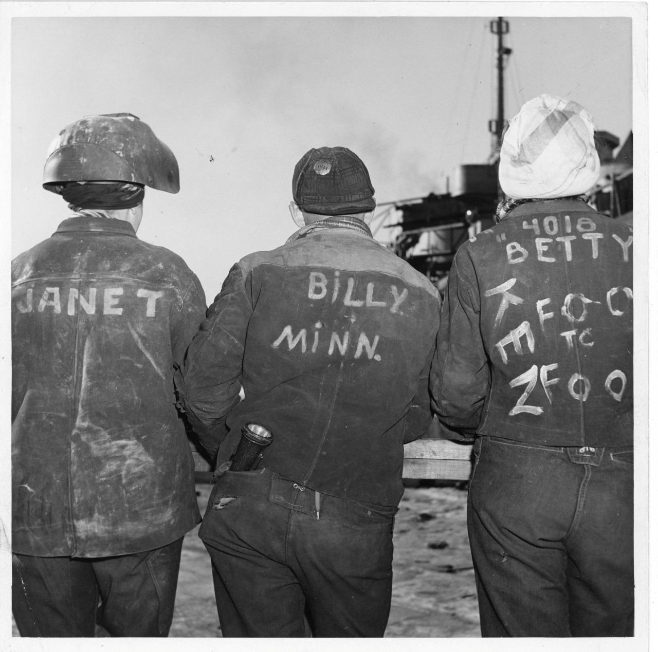 Three women in welding uniforms stand at a shipyard with their backs to the camera showing their names written on the backs of their jackets.