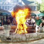 A man in a large stone-lined ring walks around a large open fire. The accoutrement of a fishboil, a boil pot, and strainer basket, plastic buckets, and a two-sided spork are at hand. A crowd of people seated at tables watch on.