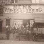 Photograph of a horse and cart and a group of men posed before a building with a large sign that reads “Henk Mineral Spring Bottling Work’s”