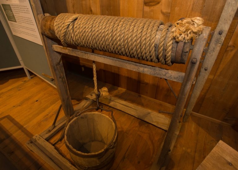 A hand-crank windlass, with a wooden frame, and a rope with a bucket on the end in a museum.