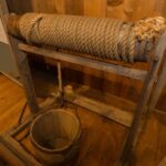 A hand-crank windlass, with a wooden frame, and a rope with a bucket on the end in a museum.