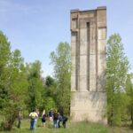 A picture of the Kickapoo Valley Reserve dam tower showing a group of people before a concrete tower rising nearly 100 feet above a grassy lawn