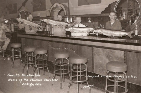 Four men pose behind a bar holding large muskie mounts. Text on photograph reads, "Suick's Muskie Bar, Home of the Muskie Thriller, Antigo, Wis. Largest Muskie display in U.S."