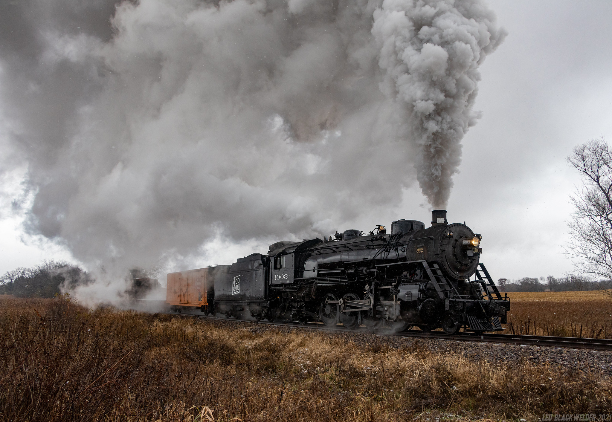 The Soo Line Engine #1003, smoke pouring out of the engine stacks, travels through an agricultural landscape.