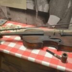 Photo of a violin and bow in a museum display, set on a table with gingham-patterned table cloth, set amongst other small items, like a pipe, a deck of cards, and a folding knife.