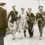 A photograph focused on four men walking down the street. Three wear gas masks and carry nightsticks. They hold another man by his arms, guiding him down the street.