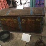 A wooden trunk, painted with blue and yellow line details resembling straps. Script text reads, "Halvor Andersen Lovans 1860"
