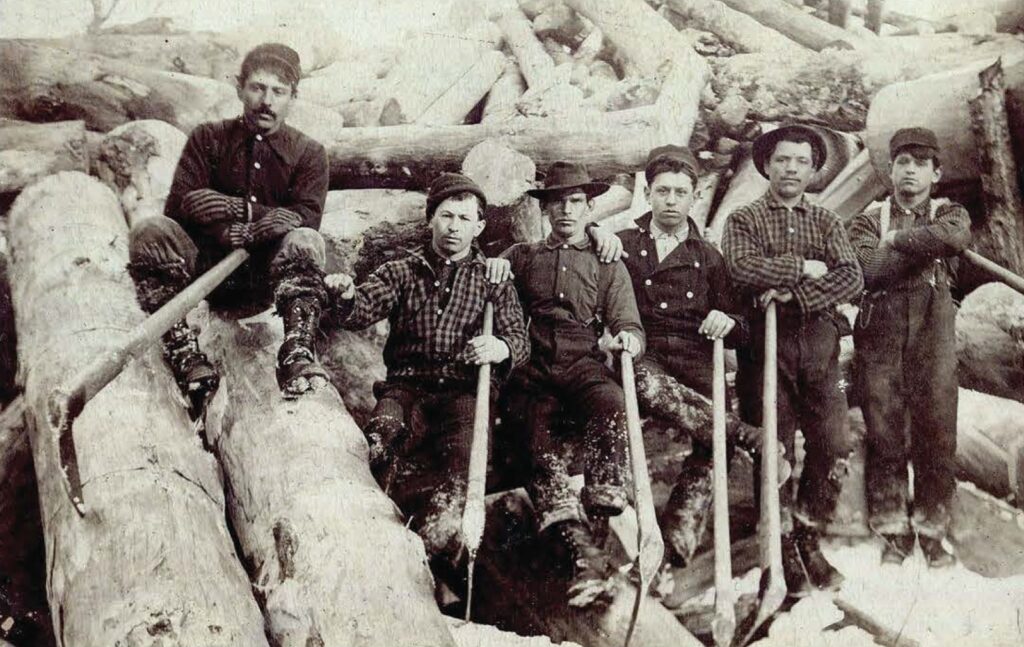 Six men pose with axes atop a pile of de-barked logs.