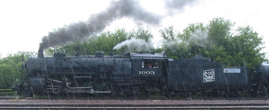 A photograph of a steam engine operating in the rain. Smoke and steam pour from its stacks.