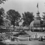 Black-and-white photograph of a spring water park, with a pool in the foreground, a pavilion in the midground, and a large hotel building in the background.