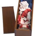 A small wooden box filled with red, white, and blue fabrics. The butt of a bottle, encased in woven red and white silk ribbons is visible at the bottom of the box.