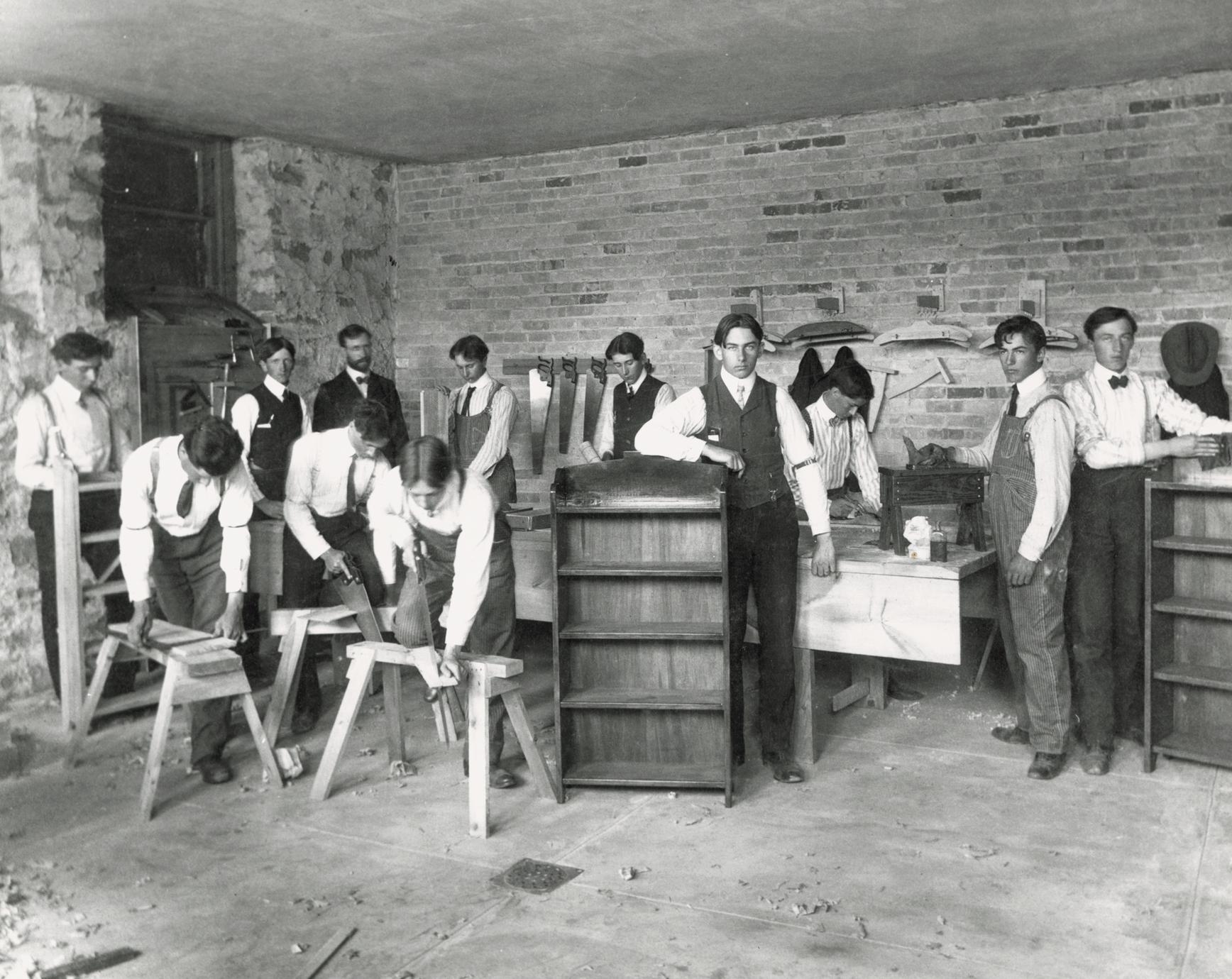 Posed image of students in a cabinetmaking class. At the center of the image a student leans against a finished cabinet. Other students are engaged in various activities like sawing and planing wood. A teacher observes in the background.
