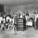 Posed image of students in a cabinetmaking class. At the center of the image a student leans against a finished cabinet. Other students are engaged in various activities like sawing and planing wood. A teacher observes in the background.